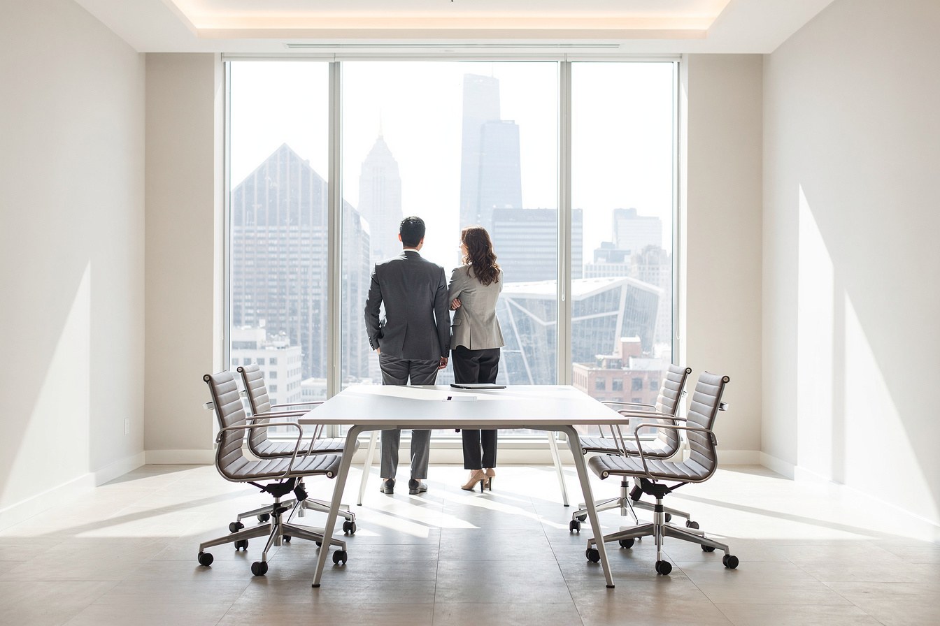 Two colleagues from behind, standing at a window discussing plans in a calm, uncluttered office space.