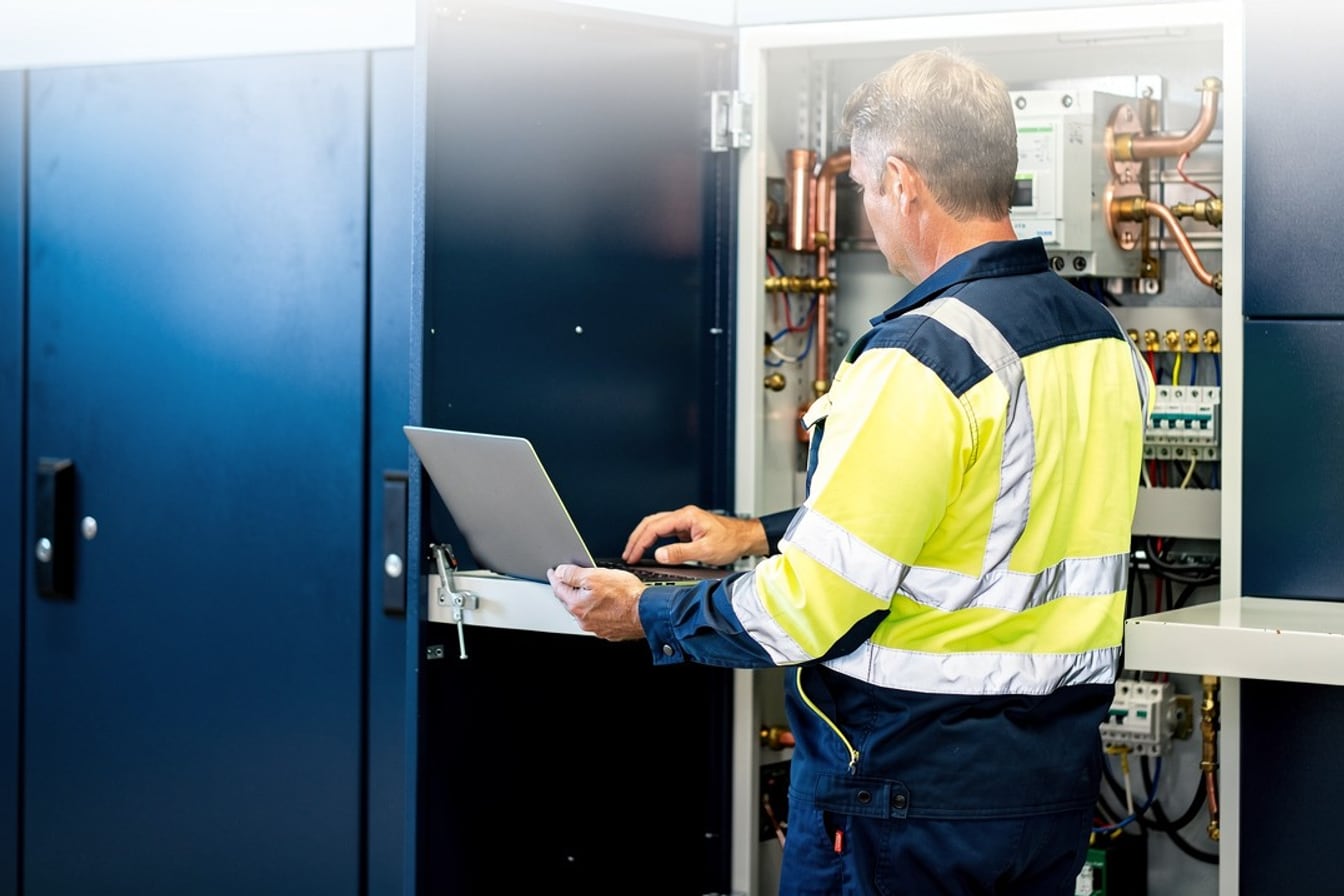 An electrician in high-vis checking a laptop beside an open switchboard on a Queensland commercial site.